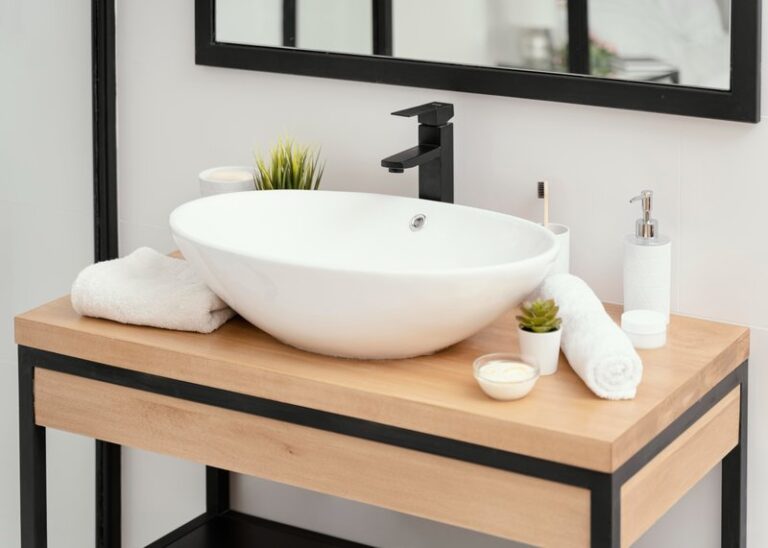 A modern bathroom sink setup with a white oval basin, black faucet, wooden countertop, white towels, a soap dispenser, a toothbrush holder, a small plant, and a cream jar.